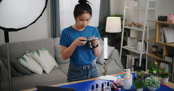 Young asian female photographer with side hustle Use laptop computer and camera, creativity her home studio as she arranges products and beauty props for photo shoot.