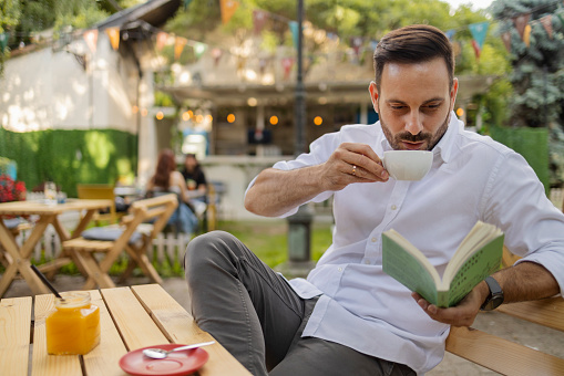 Portrait of Man Drinking Coffee and Reading a Book While Relaxing in Outdoor Cafe