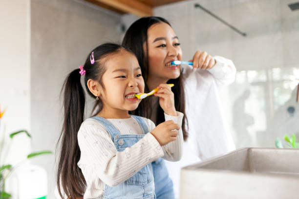 asian little girl with mom brushing teeth in bathroom, korean woman helping to brush daughter's teeth at home together - zahnpflege stock-fotos und bilder