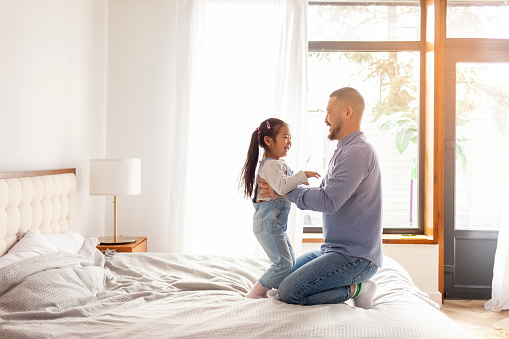 asian man dad playing with little daughter on bed at home, korean girl laughing in dad's arms, father love concept