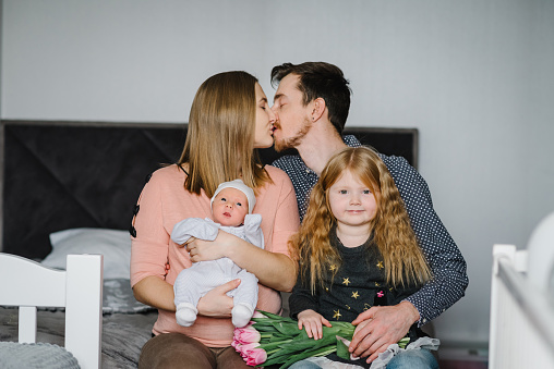 Childs, father congratulate mom with mother's day. Happy family with two children enjoying together sitting at home. Family relaxing on bed. Mother kisses dad, baby son, daughter laughing and hugging.