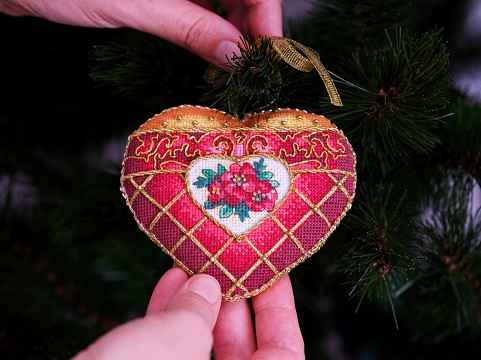 A woman hanging a red cross stitch Christmas Timeless Elegance ornament on to a Christmas tree. This heart shape Christmas ornament with red flowers embroidered and made by myself.