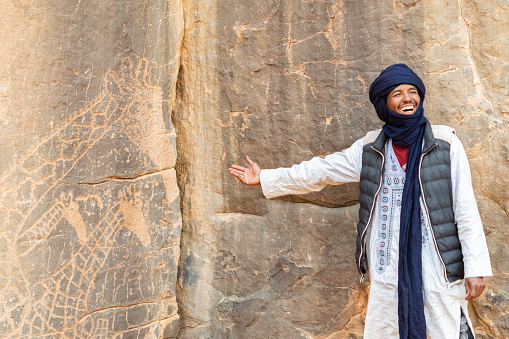 Djanet, Illizi, Algeria- December 22, 2021: a man wearing traditional clothing laughing and extending his hand towards the stone wall drawings in the Tassili N'Ajjer National Park.