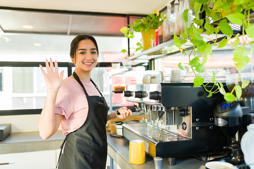 Latin happy woman and barista waving hello to a customer while smiling and working preparing coffee or cappuccino