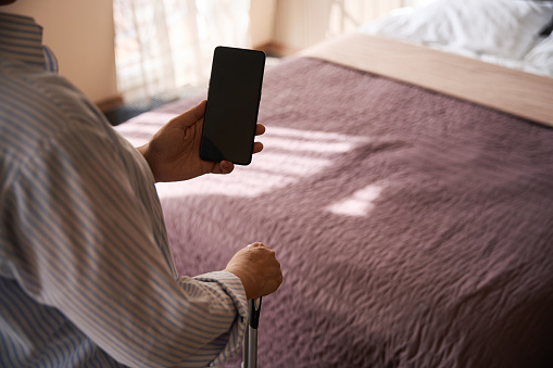 Cropped photo of lady with mobile phone in hand leaning on trolley suitcase handle in hotel room