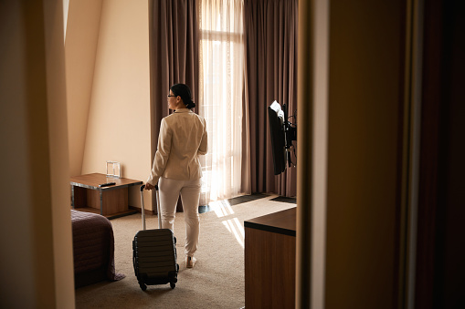 Back view of adult woman with trolley suitcase standing in middle of her suite
