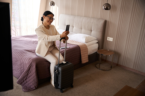 Smiling woman seated on bed in hotel room leaning on travel suitcase handle looking at cellular phone in her hand