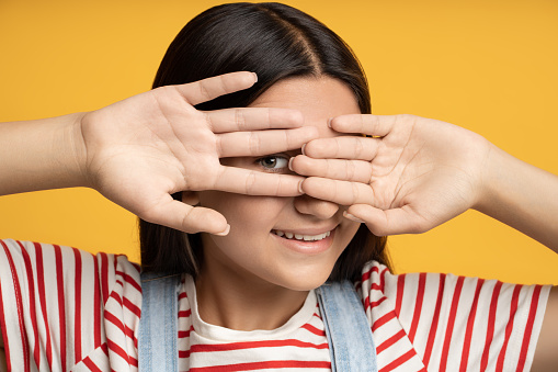 Portrait playful smiling teen girl hiding looking through hand one eye on yellow background. Brunette happy teenager playing hide and seek game. Banner, poster for advertisement, marketing concept.