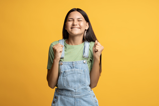 Glad smiling teenager clenching fists of great happiness, luck, having goal achievement, success, good news. Schoolgirl with closed eyes excited with positive emotions isolated on orange background