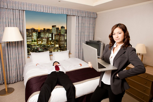 In a hotel, an exhausted young adult male businessman is laid flat out on the bed while his business partner, a young adult asian female is standing at the foot of the bed with an open laptop in one arm with an irritated expression on her face. The drapes are open, revealing a beautiful modern cityscape at sunset.