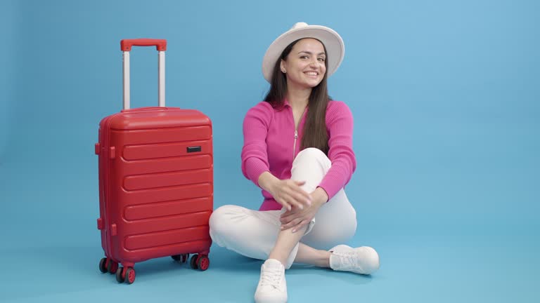 Female tourist girl is sitting and a red suitcase approaches her sign in studio on blue background. Female puts on a hat