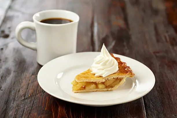 Apple pie with a whipped cream and a cup ofcoffee on rustic wooden table.  Shot wide to leave space for copy.   Professionally shot, color corrected, exported 16 bit and retouched for maximum image quality. 