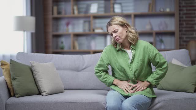 Sick mature woman feeling stomach ache while is sitting on sofa in living room at home.