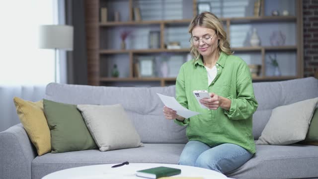 Mature woman uses mobile app for spending bills using smartphone phone while sitting at home