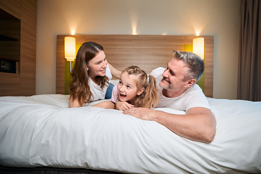 Happy caucasian family of mother, father and pleased daughter embracing while lying on bed in hotel room. Concept of rest, vacation and travelling. Family relationship and enjoying time together