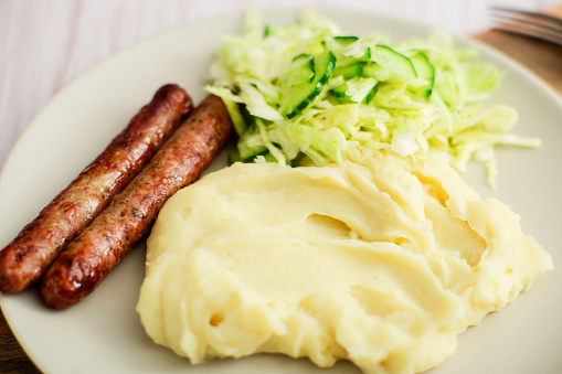 Sausages, mashed potatoes, and a zingy cabbage-cucumber salad elegantly displayed on white dinnerware against a wooden background