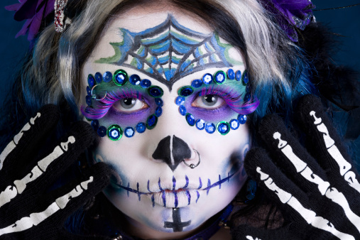 Horizontal closeup full frame studio shot of woman's face with blue sugarskull makeup and skeleton gloves.