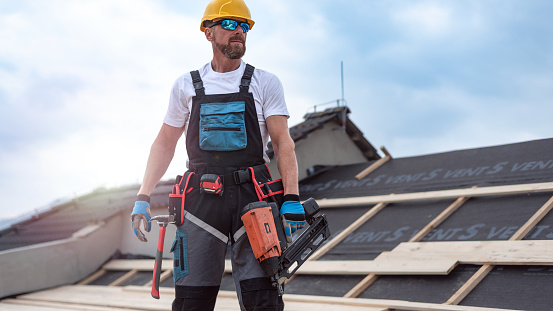 The carpenter nails a timber board using an electric nailer while working on a roof.