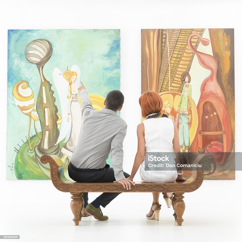 couple talking about an artwork young caucasian couple sitting on a wooden bench in an art gallery pointing at some painting Exhibition Stock Photo