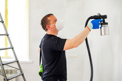 Man in protective mask paint a wall with an electric spray gun. Electric spray painting. Repair of the apartment