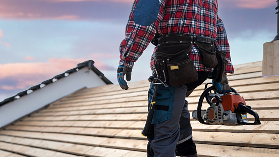 A carpenter with a tool belt holds a chainsaw in his hand while working on the roof.