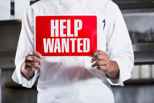 Chef in restaurant kitchen, holding Help Wanted sign.