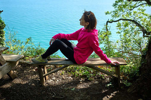 A young woman is resting while sitting in the forest on a high bank near the blue sea. Healthy lifestyle concept.