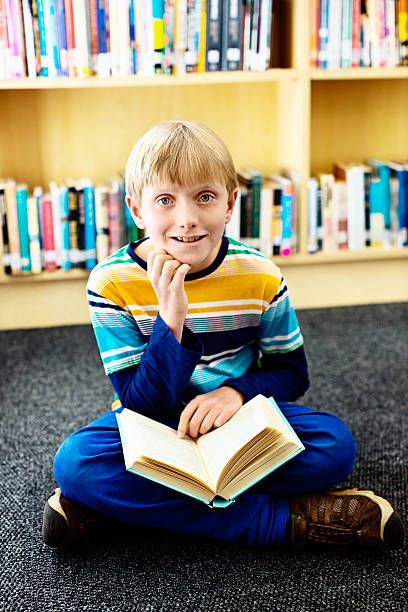 istock Smiling blond schoolboy sits on library floor reading 187307654