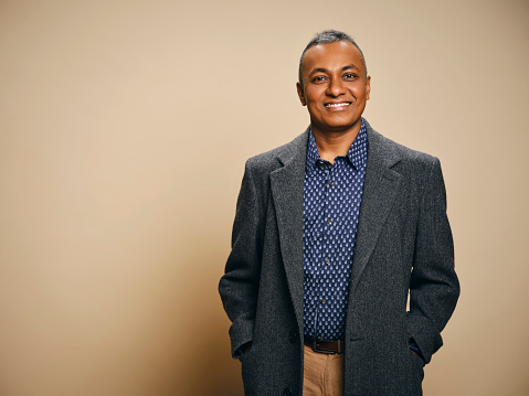 A studio portrait of a mature Indian ethnicity man, against a light brown background.