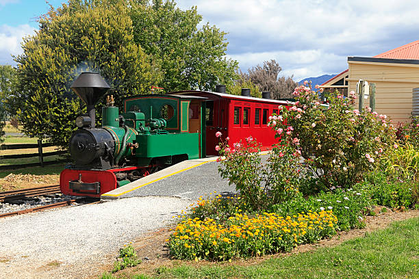 A small green vintage steam locomotive with red carriage waits at a pretty station.  In the foreground a profusion of colour from roses and other flowers.  In the background are evergreen trees. NOTE: Taken with permission of the railway owner who stabled the train in the station for the photo.  Numbers and identification marks removed. Horizontal format, copy space.