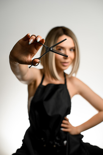 Young beautiful hairdresser woman holding scissors against face on white wall background, studio shot. Haircut coiffure haircare concept. Copy space