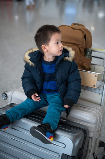 Charming two year old boy eagerly sits on suitcases in a bustling airport terminal, near check - in desk counter. Ideal for projects emphasizing family adventures and the excitement of travel with young children