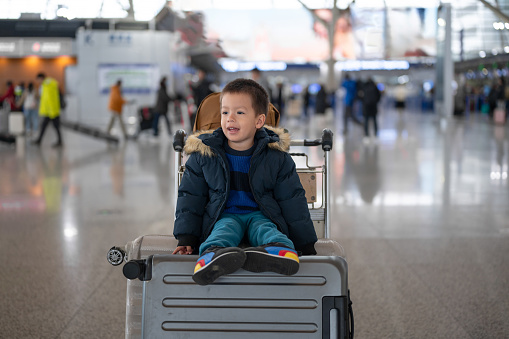 Charming two year old boy eagerly sits on suitcases and smiling in a bustling airport terminal, near check - in desk counter. Ideal for projects emphasizing family adventures and the excitement of travel with young children