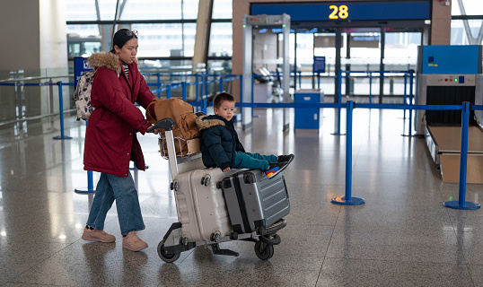 Charming two year old boy, accompanied by his Chinese mother, eagerly sits on suitcases in a bustling airport terminal, near check - in desk counter. Ideal for projects emphasizing family adventures and the excitement of travel with young children