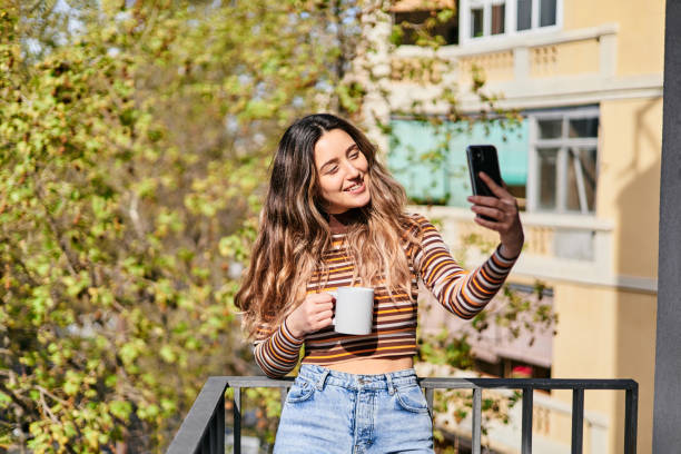 istock Cheerful woman drinking coffee while taking a selfie with her mobile phone. She is on the terrace of her house. 1873204554
