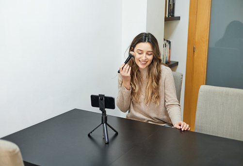 Young woman recording a video blog with her mobile phone while showing how she does her make-up.