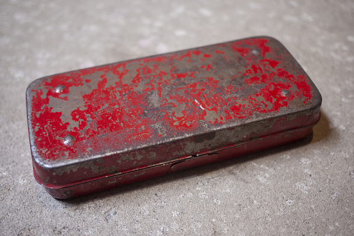 Weathered old red painted metal box on a cement floor. Small container for a a socket wrench set. Paint chipped and worn.