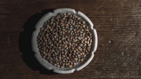 Coriander seeds, in a close-up view, rest in a bowl on a textured wooden background. The warm, earthy tones of the seeds complement the rustic charm of the setting, creating a visually pleasing and inviting image.