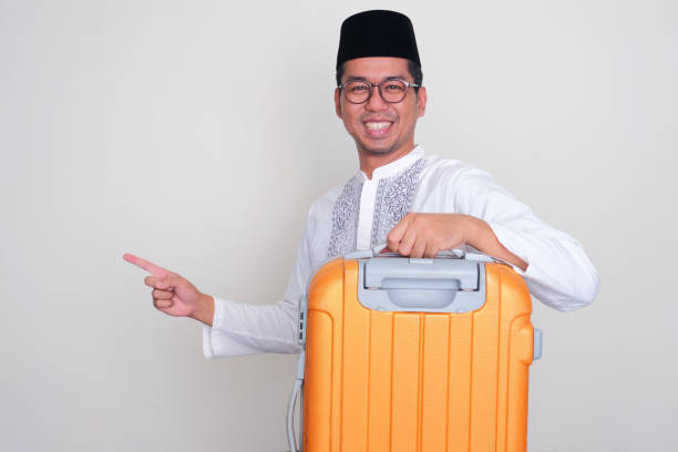 istock Moslem Asian man smiling at the camera and pointing to the right side while carrying suitcase 1873478507