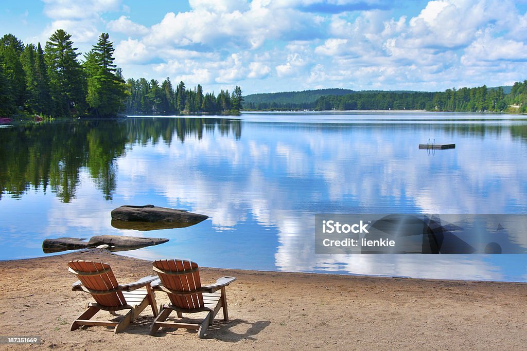 chairs lake shore two wooden deck chairs on the shore of a lake in Canada with clouds reflecting on the water Summer Stock Photo