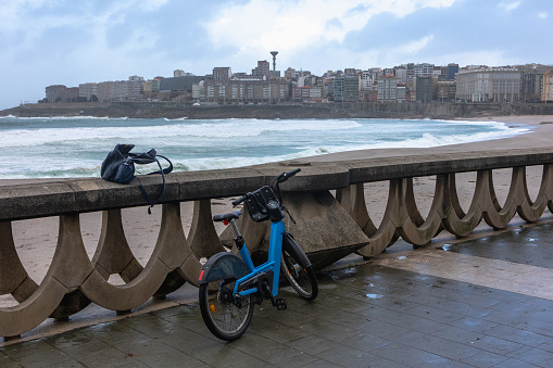 The composition captures a tranquil moment along the seaside promenade with a parked rental bike and a casually placed bag atop the balustrade, suggesting a temporary pause in someone's journey. The cityscape in the background appears hazy, possibly due to sea mist or impending rain, while the tumultuous sea churns with life and energy. This scene contrasts the stillness of the bike and bag with the dynamic nature of the ocean and city life, evoking a sense of calm amidst the everyday hustle.