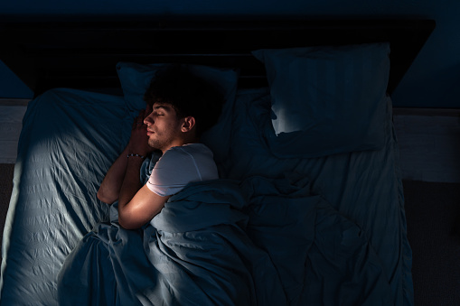 Top view of young Arabic man sleeping cozily on a comfortable bed in his bedroom at night with blue light shining through the window. Copy space