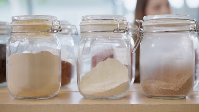 Close up shot of organic food ingredient powders stored in a glass container in a healthy refilling shop with the female customer walking in the store background. Small business providing sustainable products.