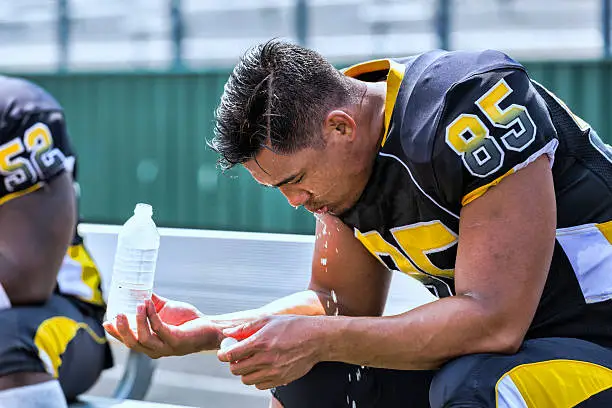 A football player sits on the bench after playing his heart out and pours cold water on himself.