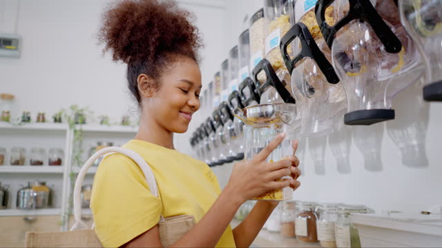 Young adult African woman wearing casual clothing choosing the yellow snack into a jar from the transparent glass container at the refilling store. Healthy female buying a healthy snack for supporting sustainable lifestyle.