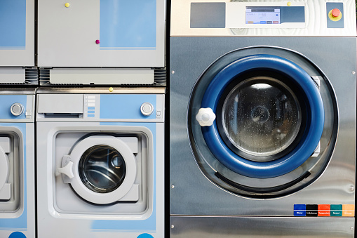 White and blue automatic washing machines standing next to bigger silver one with drum filled with clothes being cleaned with soap powder