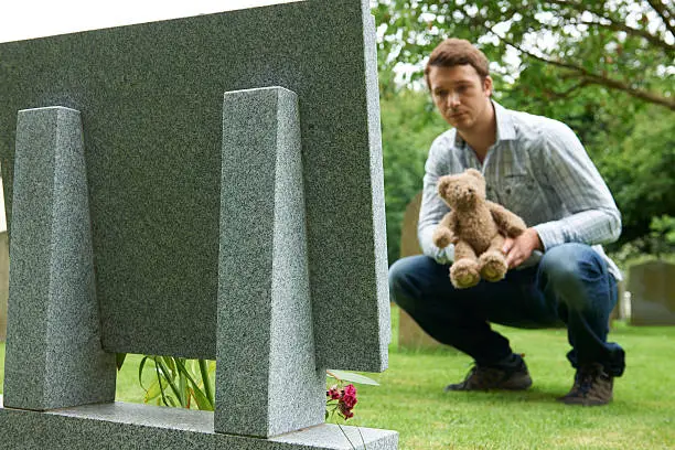 Photo of Father Placing Teddy Bear On Child's Grave In Cemetery