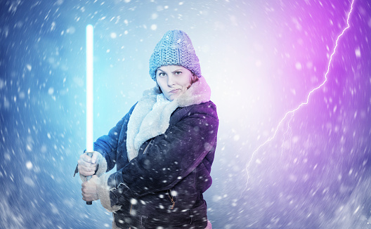 A young woman in winter clothes holds a light sword  in her hands, snow is swirling around, on a colored background.