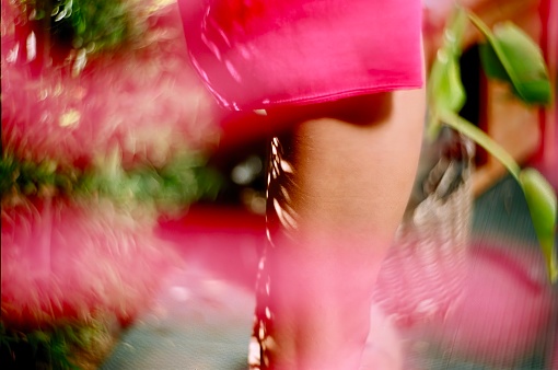 This photograph captures a blurred image of a woman in a pink dress standing near a flowering tree, creating an impressionistic effect with vibrant colors and soft focus.