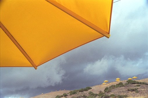 This photograph captures a vibrant yellow umbrella in the foreground with a backdrop of a stormy sky and a hillside dotted with similar umbrellas in the distance.